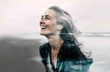 A smiling woman with wavy hair is shown in a double exposure, blending her portrait with an ocean shoreline, creating a harmonious and artistic composition.