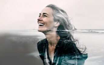 A smiling woman with wavy hair is shown in a double exposure, blending her portrait with an ocean shoreline, creating a harmonious and artistic composition.
