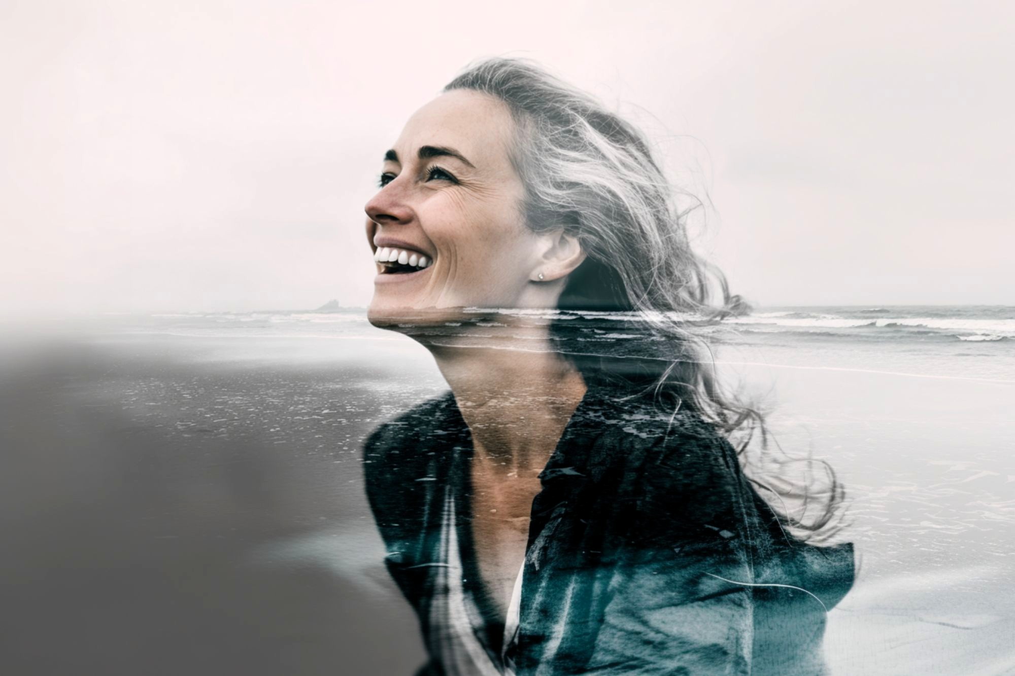 A smiling woman with wavy hair is shown in a double exposure, blending her portrait with an ocean shoreline, creating a harmonious and artistic composition.