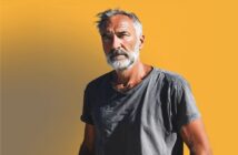 An older man with gray hair and a beard stands against a bright yellow background, wearing a wrinkled gray t-shirt and looking thoughtfully at the camera.