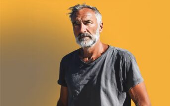 An older man with gray hair and a beard stands against a bright yellow background, wearing a wrinkled gray t-shirt and looking thoughtfully at the camera.