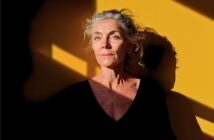 An older woman with gray hair stands against a yellow wall with sunlight and shadow patterns. She wears a black top and looks thoughtful, gazing slightly upward.