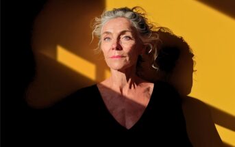 An older woman with gray hair stands against a yellow wall with sunlight and shadow patterns. She wears a black top and looks thoughtful, gazing slightly upward.