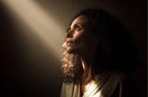 A woman with curly hair gazes upward, her face softly illuminated by a beam of light against a dark background, creating a contemplative and hopeful mood.