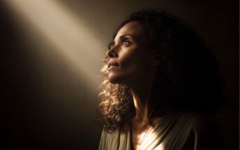 A woman with curly hair gazes upward, her face softly illuminated by a beam of light against a dark background, creating a contemplative and hopeful mood.