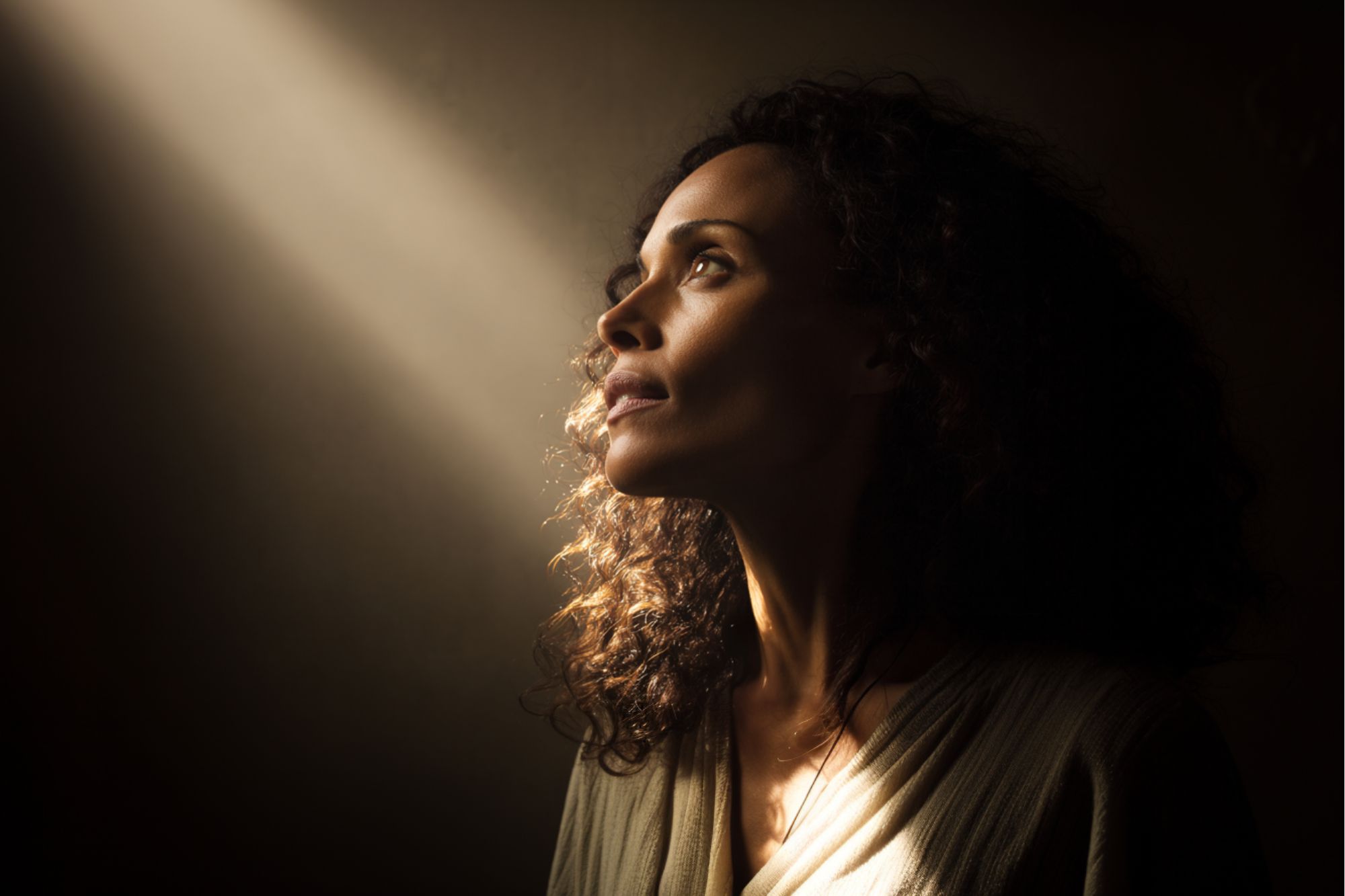 A woman with curly hair gazes upward, her face softly illuminated by a beam of light against a dark background, creating a contemplative and hopeful mood.