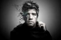 A grayscale portrait of a man with blue eyes, whose left side is dissolving into particles, creating a surreal, fragmented effect against a blurred gray background.