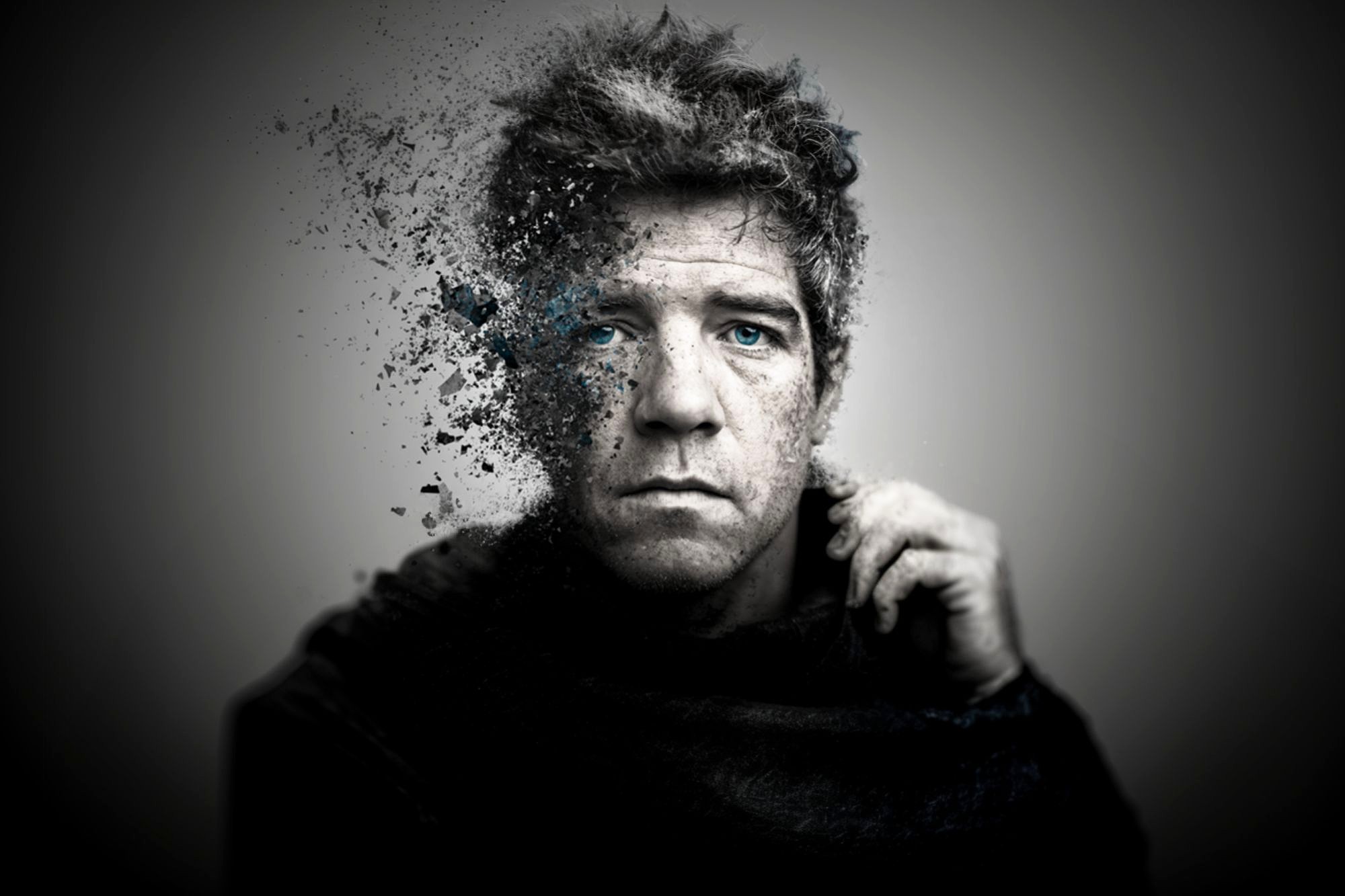 A grayscale portrait of a man with blue eyes, whose left side is dissolving into particles, creating a surreal, fragmented effect against a blurred gray background.