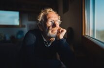 An older man with glasses and a beard rests his chin on his hand, gazing thoughtfully out of a sunlit window. He appears relaxed and contemplative, sitting indoors in a softly lit room.