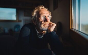 An older man with glasses and a beard rests his chin on his hand, gazing thoughtfully out of a sunlit window. He appears relaxed and contemplative, sitting indoors in a softly lit room.