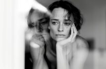 A woman with short hair rests her face on her hand, looking contemplative. Her reflection appears in a window, creating a double image effect. The photo is in black and white.