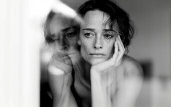 A woman with short hair rests her face on her hand, looking contemplative. Her reflection appears in a window, creating a double image effect. The photo is in black and white.