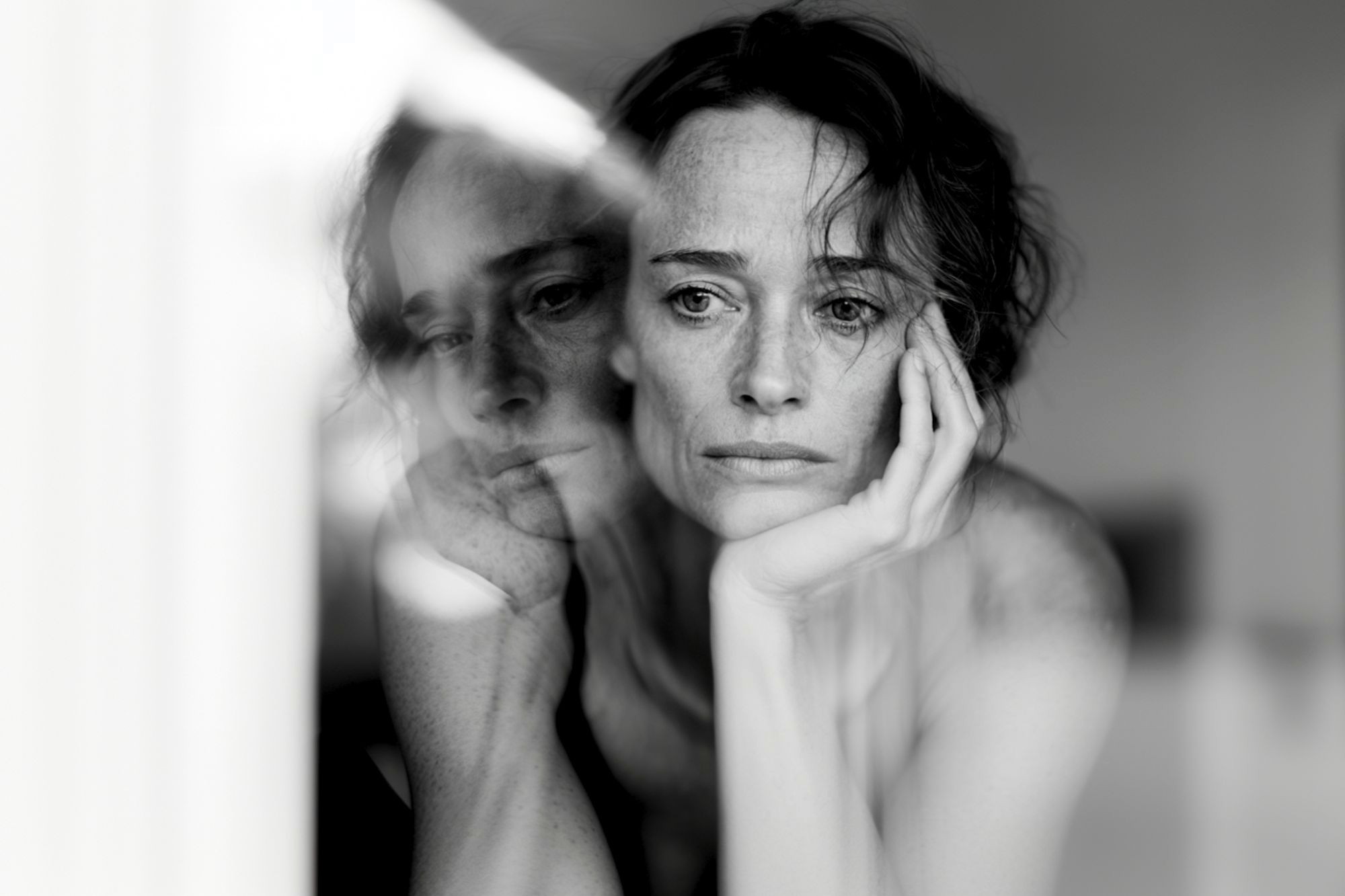 A woman with short hair rests her face on her hand, looking contemplative. Her reflection appears in a window, creating a double image effect. The photo is in black and white.