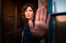 A woman with wavy brown hair stands in a doorway, facing the camera and holding up her hand as if signaling stop. Her expression is serious, and she wears a dark jacket. The background is softly blurred.
