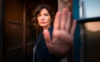 A woman with wavy brown hair stands in a doorway, facing the camera and holding up her hand as if signaling stop. Her expression is serious, and she wears a dark jacket. The background is softly blurred.