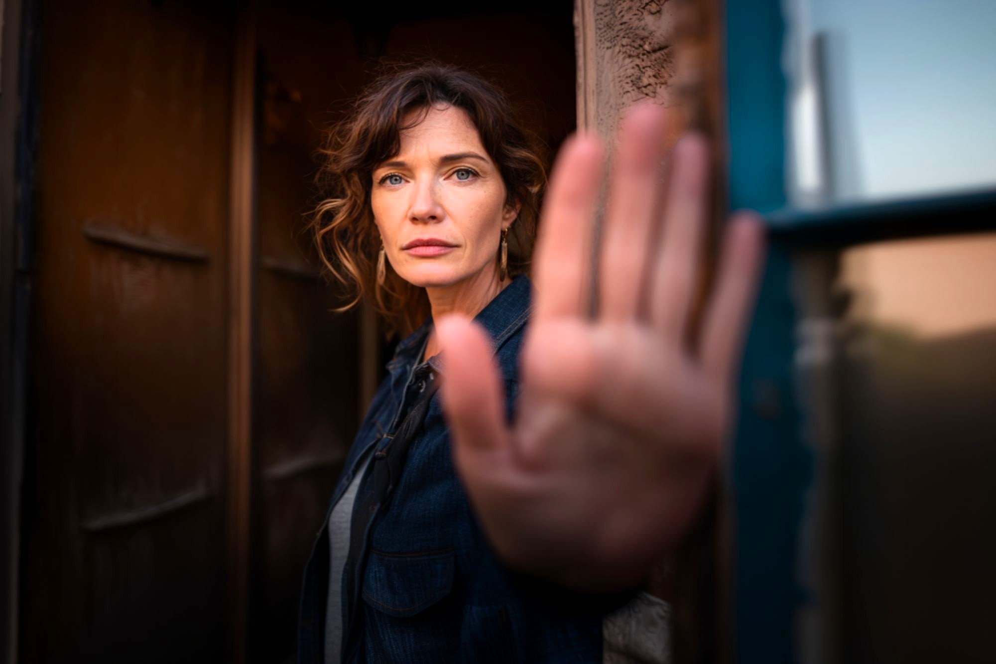 A woman with wavy brown hair stands in a doorway, facing the camera and holding up her hand as if signaling stop. Her expression is serious, and she wears a dark jacket. The background is softly blurred.