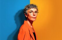 A woman with short gray hair and glasses stands against a split blue and yellow background, wearing an orange blazer and looking confidently at the camera.