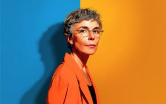 A woman with short gray hair and glasses stands against a split blue and yellow background, wearing an orange blazer and looking confidently at the camera.
