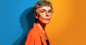 A woman with short gray hair and glasses stands against a split blue and yellow background, wearing an orange blazer and looking confidently at the camera.