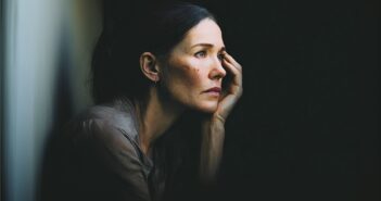 A woman with dark hair rests her face on her hand, gazing pensively out of a window with a somber expression. Soft light illuminates her face, highlighting her thoughtful mood against a dark background.