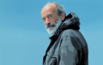 An older man with a white beard and gray hair wears a dark jacket and looks over his shoulder against a clear blue sky.