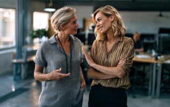 Two women stand in an office, smiling and talking to each other. One gestures with her hand while the other crosses her arms and listens. The background shows desks, chairs, and a person working at a computer.