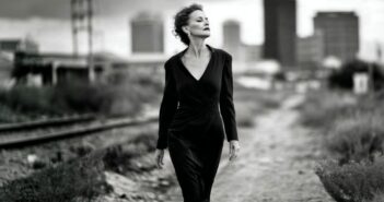 A woman in a dark dress walks confidently along a desolate path near train tracks, with a blurred city skyline in the background. The image is in black and white.