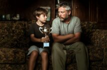 A boy holding a trophy sits on a couch next to a man with a beard. They look at each other seriously, both wearing casual clothes, in a dimly lit room.