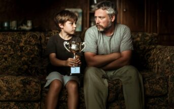 A boy holding a trophy sits on a couch next to a man with a beard. They look at each other seriously, both wearing casual clothes, in a dimly lit room.