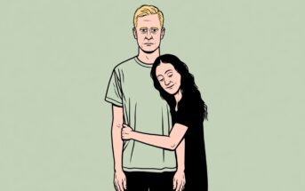 A woman with long dark hair hugs a man with short blond hair. The man stands with a neutral expression while the woman smiles with her eyes closed, leaning her head on his shoulder. The background is pale green.