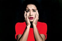 A woman in a bright red top holds her face in both hands, looking shocked or scared, with wide eyes and parted lips, set against a dark background.