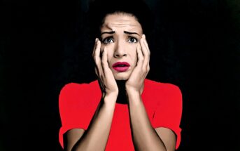 A woman in a bright red top holds her face in both hands, looking shocked or scared, with wide eyes and parted lips, set against a dark background.