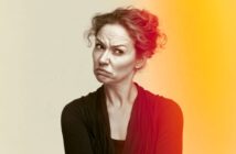 A woman with curly hair makes a skeptical facial expression. She wears a dark top, stands against a beige background, and is partially lit by an orange-yellow light on the right side of the image.