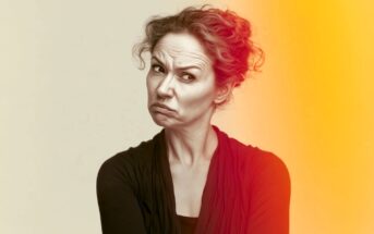 A woman with curly hair makes a skeptical facial expression. She wears a dark top, stands against a beige background, and is partially lit by an orange-yellow light on the right side of the image.