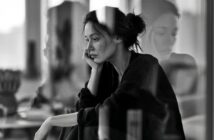 A woman sits by a window with her head resting on her hand, looking pensive. Her reflection is visible in the glass. The image is in black and white, creating a contemplative and moody atmosphere.