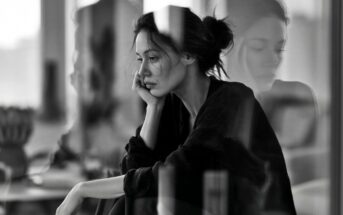 A woman sits by a window with her head resting on her hand, looking pensive. Her reflection is visible in the glass. The image is in black and white, creating a contemplative and moody atmosphere.