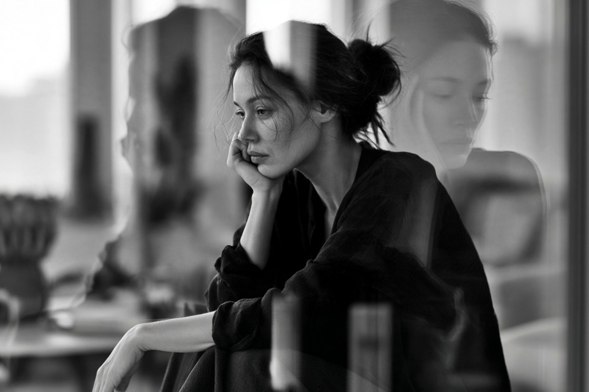 A woman sits by a window with her head resting on her hand, looking pensive. Her reflection is visible in the glass. The image is in black and white, creating a contemplative and moody atmosphere.