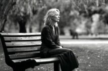 A woman with light hair sits alone on a park bench, looking thoughtful. The black-and-white photo shows trees and blurred benches in the background, suggesting a calm, quiet park setting.