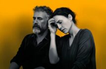 A man and woman sit close together against a bright yellow background. The woman rests her head on the man’s shoulder, eyes closed, while the man looks serious and thoughtful. Both appear somber and deep in thought.