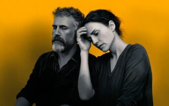 A man and woman sit close together against a bright yellow background. The woman rests her head on the man’s shoulder, eyes closed, while the man looks serious and thoughtful. Both appear somber and deep in thought.