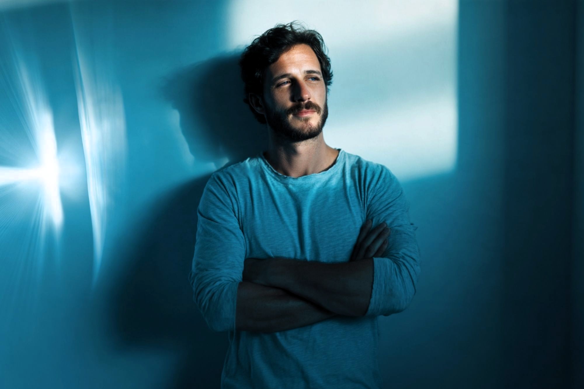 A man with short hair and a beard stands against a blue-lit wall with his arms crossed, looking thoughtful. Light and shadows create an artistic effect on the wall behind him.