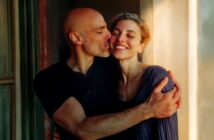 A smiling woman with curly hair is hugged and kissed on the cheek by a bald man near a sunlit window. Both appear happy and affectionate.