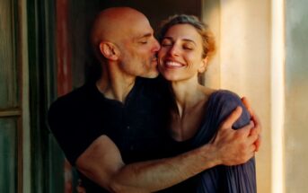 A smiling woman with curly hair is hugged and kissed on the cheek by a bald man near a sunlit window. Both appear happy and affectionate.