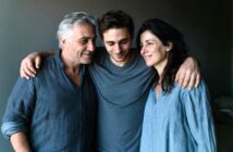 Three people stand closely together, smiling. A young man in the center has his arms around an older man and woman on either side. All wear casual blue tops and appear happy and relaxed.