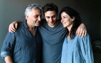 Three people stand closely together, smiling. A young man in the center has his arms around an older man and woman on either side. All wear casual blue tops and appear happy and relaxed.