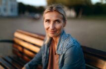 A woman with blonde hair and blue eyes sits on a wooden bench outdoors, smiling gently at the camera. She wears a denim jacket and neutral top, with a soft background of trees and buildings.