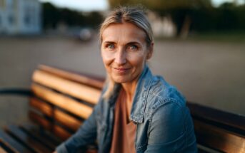A woman with blonde hair and blue eyes sits on a wooden bench outdoors, smiling gently at the camera. She wears a denim jacket and neutral top, with a soft background of trees and buildings.