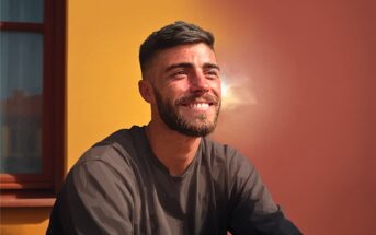 A man with short dark hair and a beard smiles while sitting in sunlight, in front of a wall that is half yellow and half reddish-brown, near a window.