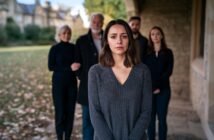 A woman stands in the foreground looking sad, while four people stand behind her outside a stone building, with autumn leaves on the ground.