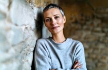 A person with short gray hair and a gray sweater stands with arms crossed, leaning against a stone wall, looking calmly at the camera.